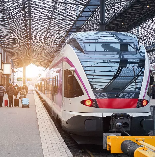 Un moderno treno in arrivo in una stazione con una struttura di vetro, passeggeri in attesa e bagagli sul marciapiede.
