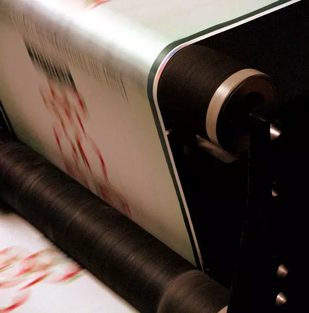 Immagine che mostra un processo di stampa con una bobina di carta che scorre attraverso una pressa. La carta presenta colori vivaci e un design grafico, evidenziando attività tipografica.
